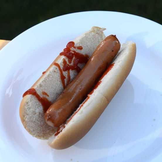 Ein weißer Teller enthält einen Hot Dog mit Brötchen, überzogen mit Ketchup. Der Hot Dog ist teilweise aufgeschnitten und zeigt das Innere.