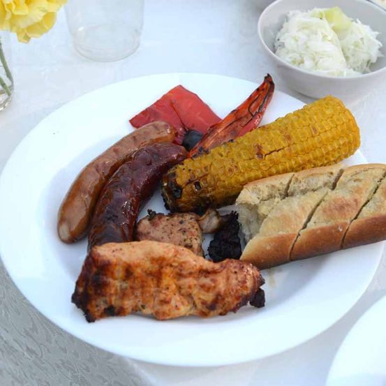 Ein weißer Teller mit gegrillten Würsten, Hähnchen, Mais, Paprika und geschnittenem Brot, mit einer Schüssel Krautsalat im Hintergrund.
