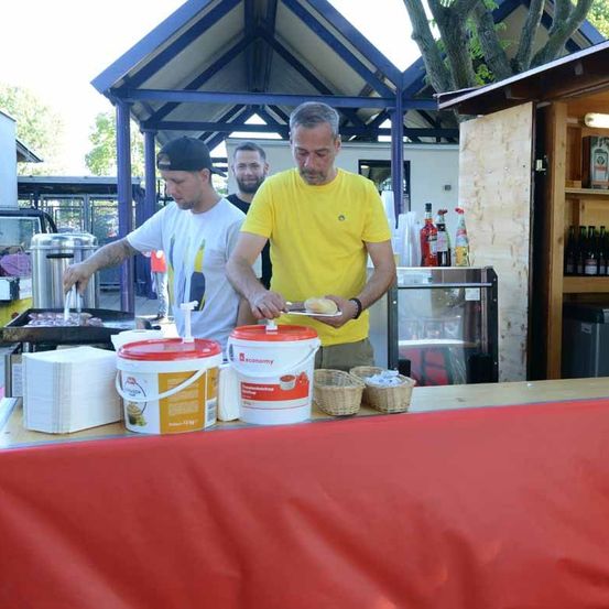 Zwei Männer bereiten Essen an einem Imbissstand vor. Ein Mann trägt ein gelbes T-Shirt und hält einen Teller mit Essen. Ein anderer Mann trägt ein weißes T-Shirt und benutzt eine Kaffeemaschine.