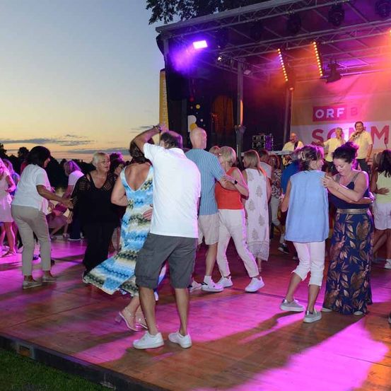 Eine Gruppe von Menschen tanzt auf einer Bühne mit einer Band vor ihnen. Die Bühne ist von rosa und lila Lichtern beleuchtet.
