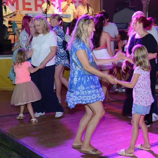 Ein Outdoor-Event mit Bühne, Frauen und Kinder tanzen, während eine Band auf der Bühne spielt.