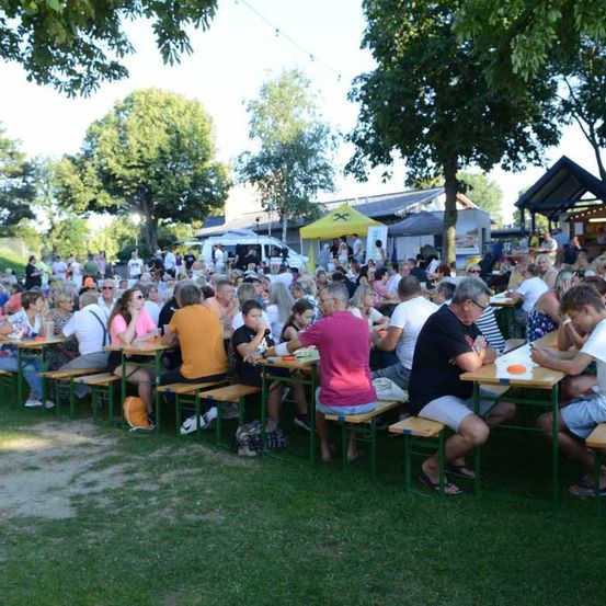 Eine große Gruppe von Menschen sitzt an Tischen auf einer Rasenfläche, mit Bäumen und einem gelben Zelt im Hintergrund. Sie genießen Essen und Getränke.