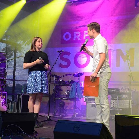 Eine Frau mit einem Mikrofon steht neben einem Mann, der einen Ordner und ein Mikrofon in der Hand hält, auf der Bühne. Hinter ihnen befindet sich ein Hintergrund mit dem Text 'ORF SOIM'. Hinter den Lautsprechern sind Musikinstrumente aufgestellt.