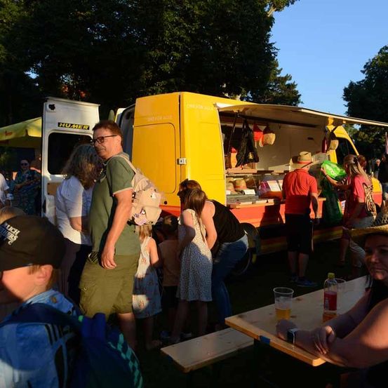 In einem Park ist ein gelber Imbisswagen geöffnet. Kunden stehen herum, einige halten Getränke. Ein Mann mit Brille und Rucksack schaut zum Wagen.