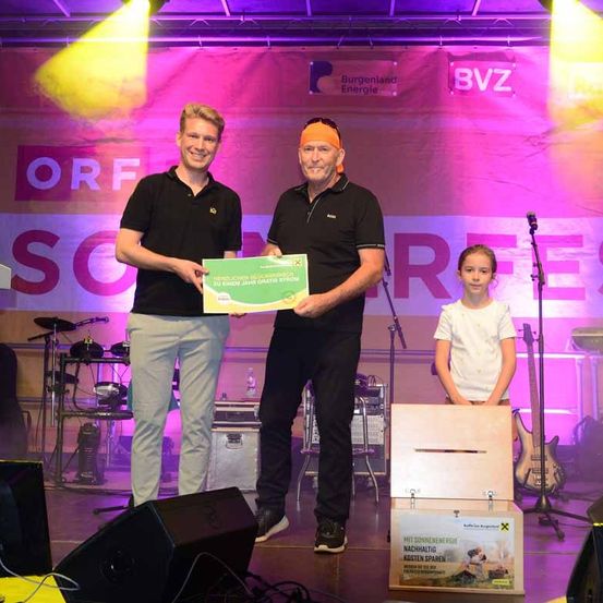 Drei Personen stehen auf einer Bühne und halten ein Zertifikat. Ein Mann in der Mitte lächelt und hält ein Zertifikat. Auf der linken Seite steht ein Mann und lächelt, und auf der rechten Seite steht ein Mädchen und lächelt. Eine Holzkiste steht auf der Bühne, und ein Mikrofon steht auf einem Stativ. Musikinstrumente stehen auf der Bühne.