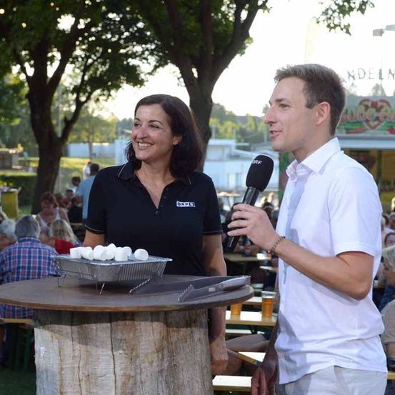 Eine Frau in einem schwarzen Shirt hält eine Schale mit Marshmallows, während ein Mann in einem weißen Shirt in ein Mikrofon spricht. Sie befinden sich in einer Outdoor-Umgebung mit Bäumen und sitzenden Menschen in der Umgebung.