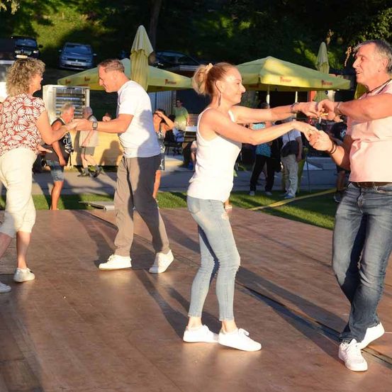 Eine Gruppe von Menschen tanzt zusammen auf einem Holzboden bei einer Veranstaltung im Freien. Die Leute sind lässig gekleidet und halten sich an den Händen. Hinter ihnen stehen Zelte und geparkte Autos.