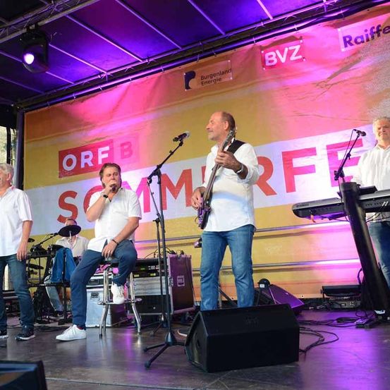 Eine Band tritt auf der Bühne auf, mit einem Banner dahinter. Ein Mitglied singt, während ein anderes Gitarre spielt.