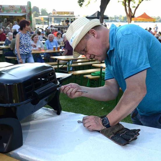 Ein Mann mit Hut und Handschuhen kocht auf einem schwarzen Grill im Freien. Hinter ihm sitzen mehrere Personen an Tischen, einige mit Getränken. Im Hintergrund befinden sich Essensbuden und ein Gewässer.