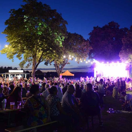 Ein nächtliches Open-Air-Konzert mit einer großen Menschenmenge, leuchtenden Lichtern und einer Bühne mit einem orangefarbenen Zelt im Hintergrund.