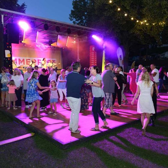 Ein nächtliches Festival mit einer Freiluftbühne. Leute tanzen auf der Bühne unter lila Lichtern. Einige Leute stehen herum und schauen zu.