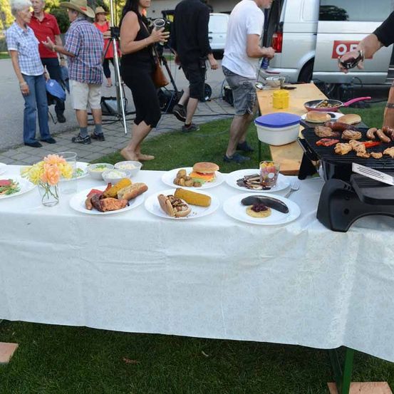 Menschen versammeln sich um einen Tisch, der mit Essen, einem Grill und einem Van im Hintergrund bedeckt ist.