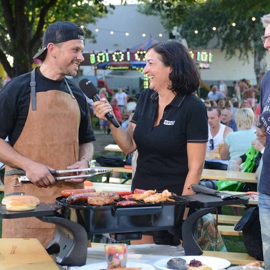 Ein Mann und eine Frau grillen bei einer Outdoor-Veranstaltung. Der Mann hält ein Mikrofon und spricht mit der Frau, die lächelt und ein Handy in der Hand hält. Der Grill steht auf einem Tisch mit Essen, und die Leute sitzen an Picknicktischen. Im Hintergrund gibt es einen Jahrmarkt und eine große Menschenmenge.