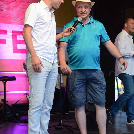 Ein Mann in einem weißen Hemd steht auf der Bühne, während ein anderer Mann in einem blauen Hemd und einem weißen Hut ein Mikrofon hält. Dahinter befindet sich eine Bühne mit einem großen roten Buchstaben E.