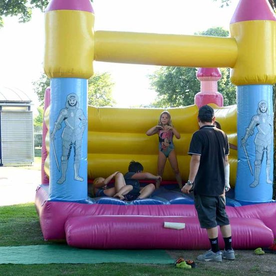 Ein bunter aufblasbarer Hüpfburg steht auf einem Rasenfeld. Eine Mädchen und zwei Kinder sind drinnen, während ein Mann draußen steht. Die Hüpfburg hat Ritterfiguren darauf.