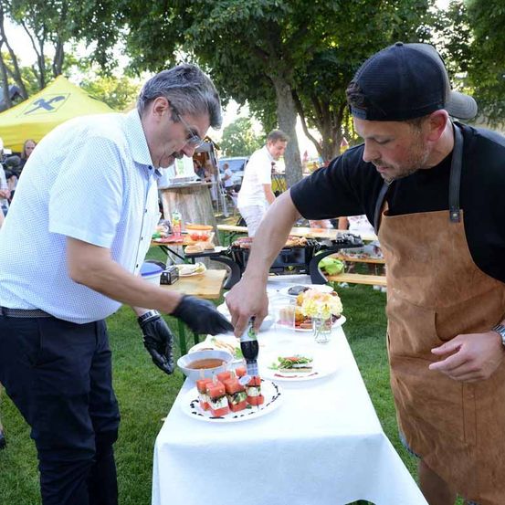 Zwei Männer bereiten auf einem Tisch bei einer Outdoor-Veranstaltung Essen zu. Einer trägt eine Schürze und der andere hat Handschuhe an.