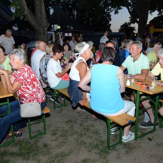 Eine Zusammenkunft älterer Erwachsener, die an Bänken unter Bäumen sitzen, einige in Freizeitkleidung, die sich unterhalten und Gläser halten.