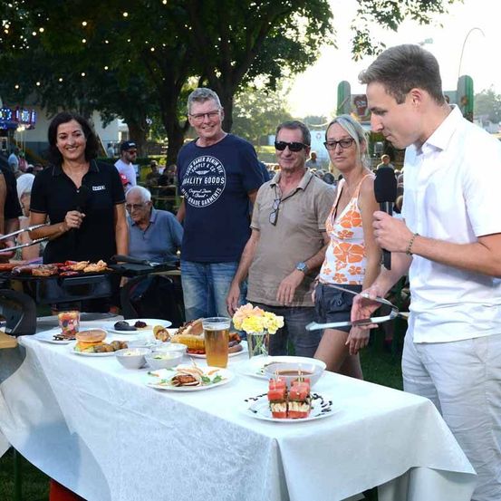 Eine Gruppe von Menschen ist bei einer Veranstaltung im Freien. Ein Mann grillt Essen, während andere das Essen und die Getränke auf dem Tisch genießen. Ein Mann hält ein Mikrofon.
