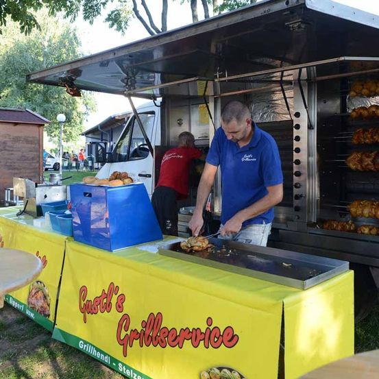 Ein Mann schneidet Essen auf einem Grill vor einem Food-Truck. Der Truck trägt den Namen 'Castl's Grillservice'. Im Hintergrund sind andere Leute zu sehen.