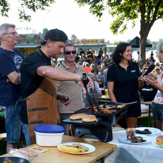 Eine Grillstation im Freien mit gegrilltem Essen. Ein Mann in einer braunen Schürze kocht, während eine Frau lächelt. Im Hintergrund stehen mehrere Personen, die zuschauen und das Ereignis genießen.