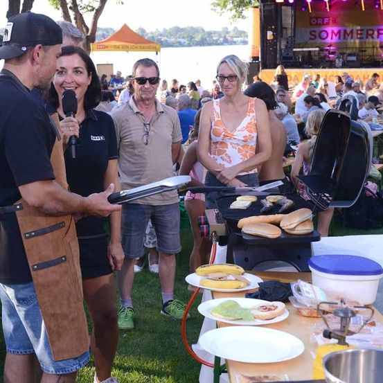 Eine Gruppe von Menschen grillt im Freien. Ein Mann hält eine Pfanne und spricht in ein Mikrofon. Hinter ihm hält eine Frau ein Mikrofon. Im Hintergrund befindet sich eine Bühne mit einem Banner und einer Menge.
