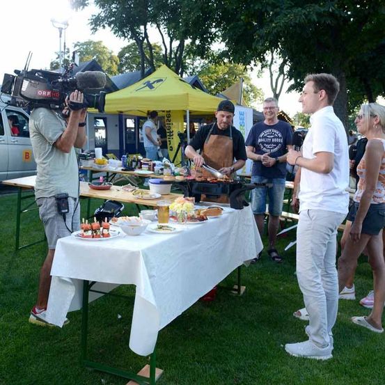Ein Filmteam filmt einen Lebensmittelverkäufer bei einer Outdoor-Veranstaltung. Der Verkäufer grillt Lebensmittel, während andere in der Nähe sprechen. Zelte, Tische und ein Van sind im Hintergrund zu sehen.