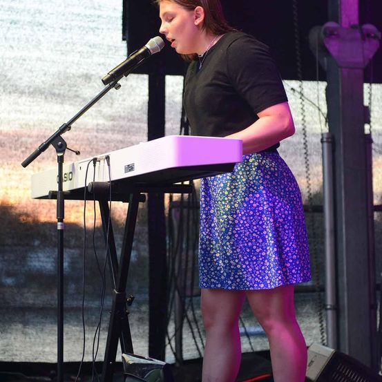 Eine Frau steht auf der Bühne und singt in ein Mikrofon. Sie trägt ein schwarzes Shirt und eine lila gemusterte Rock. Vor ihr steht ein Keyboard mit Ständer. Der Hintergrund zeigt eine verschwommene Ansicht einer Bühne mit Beleuchtung.