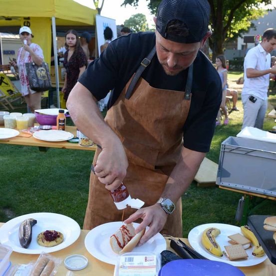 Ein Mann in einer braunen Schürze bereitet draußen auf einem Tisch Essen zu. Er hat einen Teller mit einem Hot Dog und anderen Speisen. Einige Leute stehen herum, andere sitzen. Es gibt auch andere Tische mit Essen und Getränken. In der Ferne sind Bäume und ein Zelt.