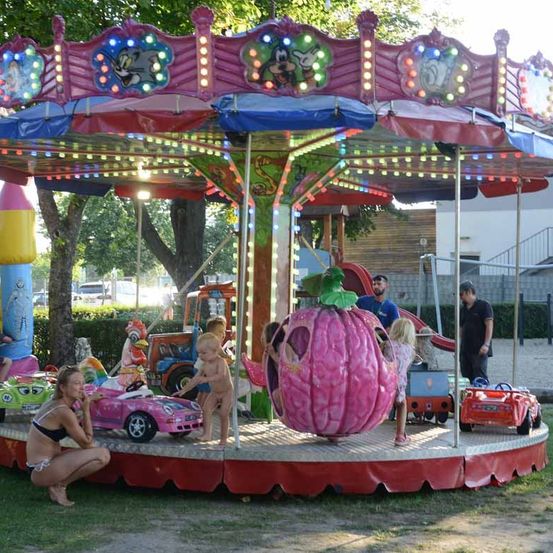 Kinder genießen eine Karussellfahrt in einem Outdoor-Vergnügungspark. Einige Kinder sitzen auf Spielzeugautos, während eine Frau auf dem Gras kniet.