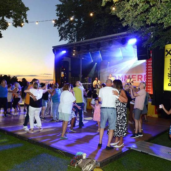 Eine Outdoor-Bühne mit tanzenden Menschen unter Lichterketten und einem Sonnenuntergang. Zwei Gitarristen spielen auf der Bühne mit einer Leinwand im Hintergrund.