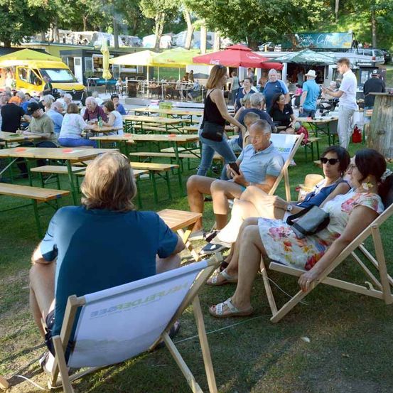 Eine Outdoor-Versammlung mit Tischen und Stühlen unter Schirmen. Eine Frau geht, während andere sitzen und reden. Fahrzeuge und Zelte im Hintergrund.