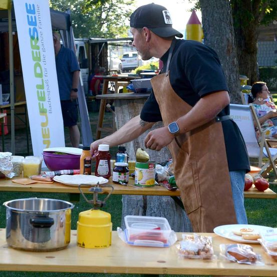 Ein Mann kocht im Freien mit einem Gasherd und hält eine Flasche Ketchup. Verschiedene Gewürze, Teller und ein Metalltopf stehen auf dem Tisch. Ein Banner mit dem Text 'EUFELDERS' ist im Hintergrund.