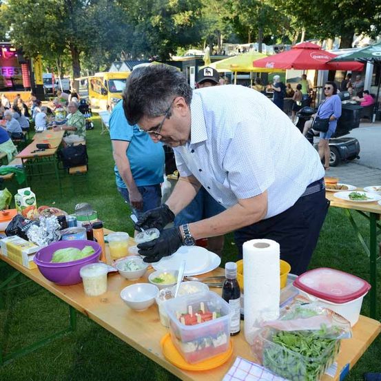 Eine Outdoor-Veranstaltung mit Essen, das auf einem Tisch zubereitet wird, mit verschiedenen Gemüsesorten und Gewürzen. Die Leute sitzen um die Tische herum. Eine Bühne mit Lautsprecher ist im Hintergrund zu sehen.