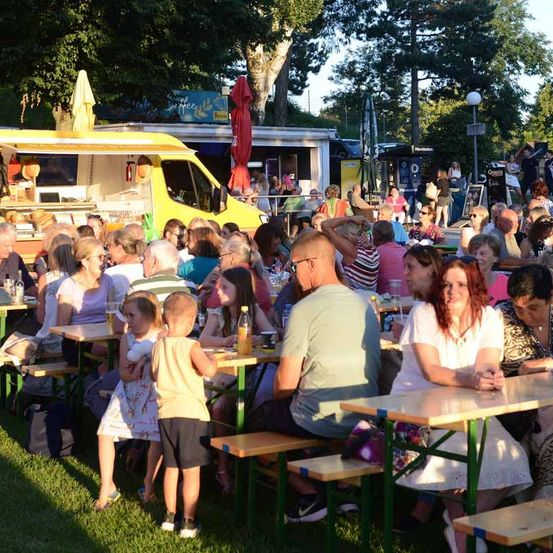 Ein Outdoor-Event mit Menschen an Tischen, Food Trucks und Bäumen im Hintergrund. Einige Personen halten Getränke und tragen Sonnenbrillen.