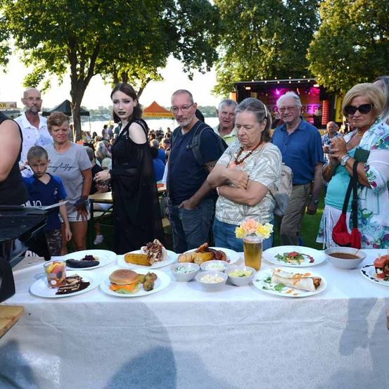 Eine Gruppe von Menschen steht um einen Tisch herum, der mit verschiedenen Speisen gefüllt ist. Der Tisch ist mit einem weißen Tuch bedeckt. Es gibt Sandwiches, Kuchen und andere Gerichte. Einige Leute schauen auf den Tisch, während andere sprechen. Im Hintergrund befinden sich Bäume und eine Bühne mit sitzenden Personen.