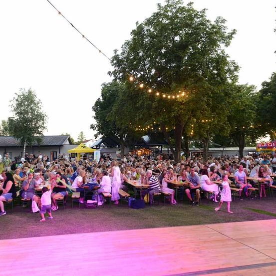 Eine große Menschenmenge versammelt sich für eine Outdoor-Veranstaltung, um Tische herumgesessen mit Lichterketten darüber. Bäume und Gebäude sind im Hintergrund sichtbar.