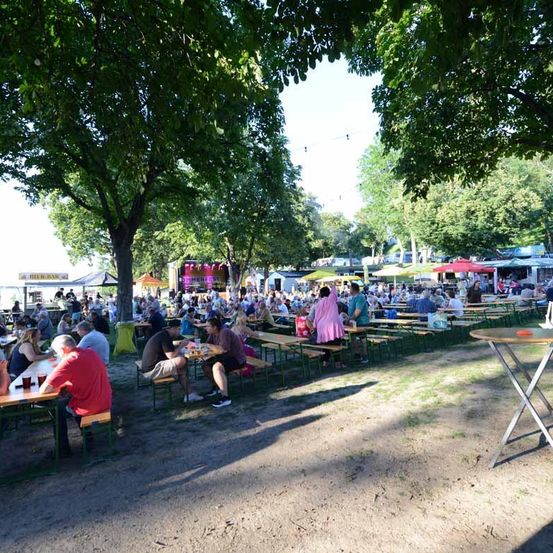 Ein lebhaftes Open-Air-Festival mit Menschen unter Sonnenschirmen, einige mit Getränken, in einem Bereich mit Bäumen und einer Bühne mit Lautsprecher-Setup.