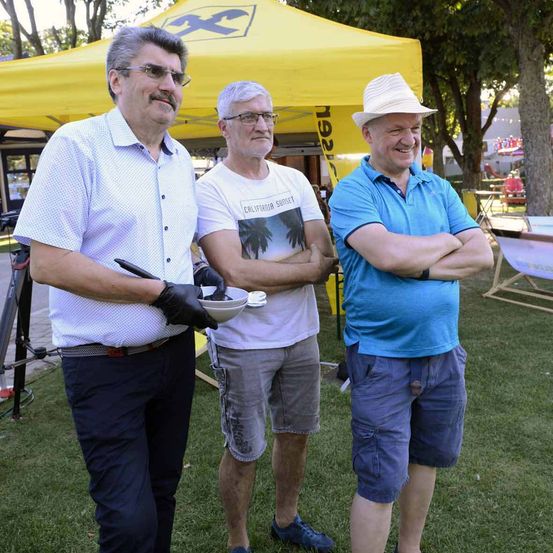 Drei Männer stehen auf einer Wiese, einer hält eine Schüssel, einer lächelt. Hinter ihnen steht ein gelbes Zelt mit einem 'X'-Logo.