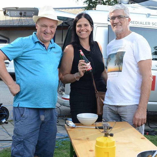 Drei Menschen stehen draußen in der Nähe eines Tisches, einer hält eine Flasche. Ein Van ist hinter ihnen geparkt.