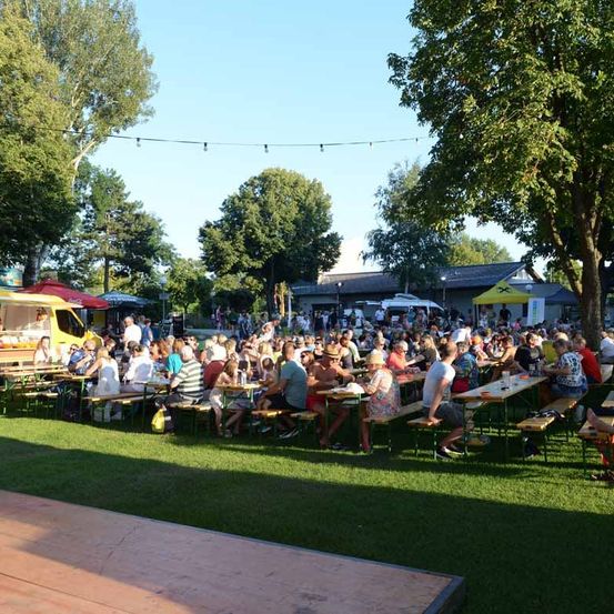 Eine große Menschenmenge versammelt sich unter Zelten in einem Park, mit Food Trucks und Bänken als Sitzgelegenheiten. Bäume und ein Gebäude sind im Hintergrund zu sehen.