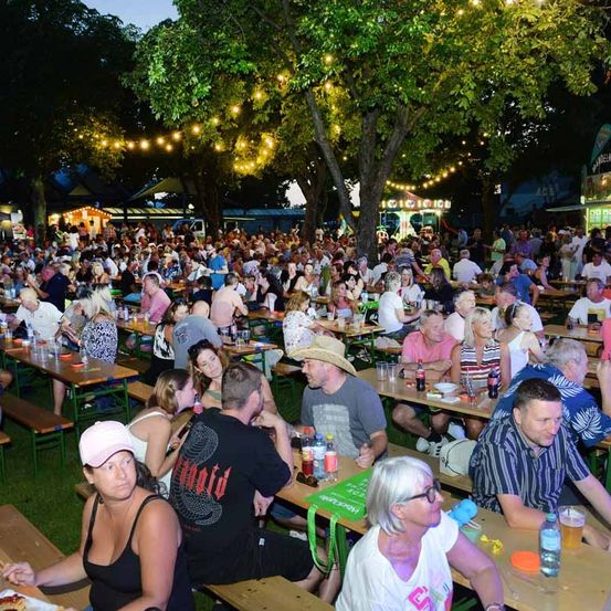 Eine große Menschenmenge sitzt bei Nacht in einem Park unter beleuchteten Bäumen an Tischen und genießt Essen und Getränke.