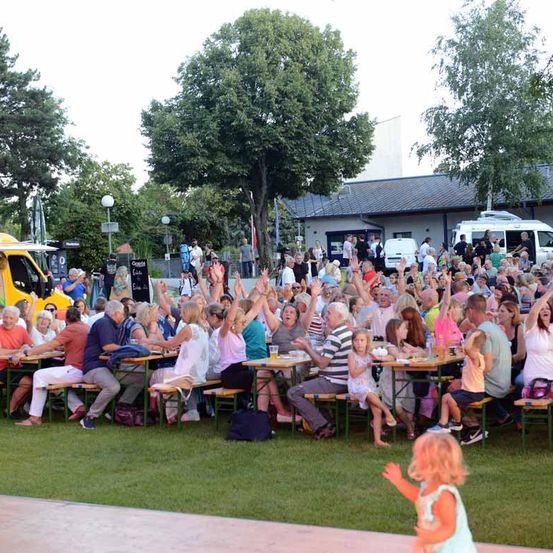 Eine Gruppe von Menschen versammelt sich auf einem Grasfeld im Freien, wahrscheinlich für eine Outdoor-Veranstaltung. Sie sitzen auf Bänken und Tischen, einige klatschen mit den Händen. Ein gelber Lastwagen ist in der Nähe geparkt. Bäume und ein Gebäude sind im Hintergrund.