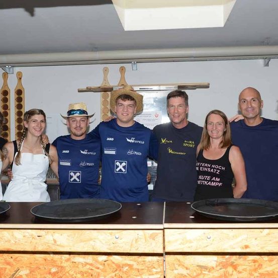 Eine Gruppe von Menschen in einem Raum, die passende blaue T-Shirts tragen, zwei Frauen in weißen Schürzen, ein Mann mit Hut und ein Schild an der Wand.