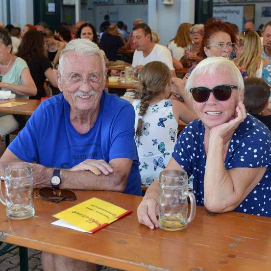 Zwei ältere Menschen sitzen an einem Tisch mit Bierkrügen in einem Outdoor-Essbereich. Sie lächeln und scheinen ihre Mahlzeit zu genießen.