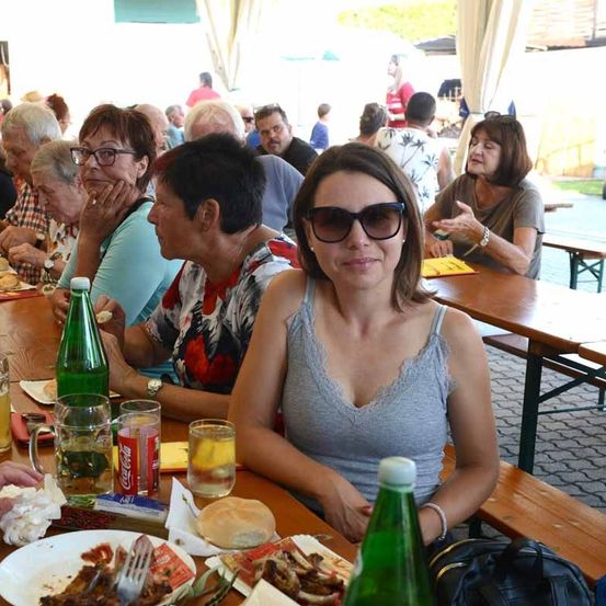 Eine Gruppe von Menschen sitzt an einem langen Tisch in einem Outdoor-Essbereich, mit Essen und Getränken vor ihnen. Eine Frau trägt eine Sonnenbrille und ein graues Tanktop.