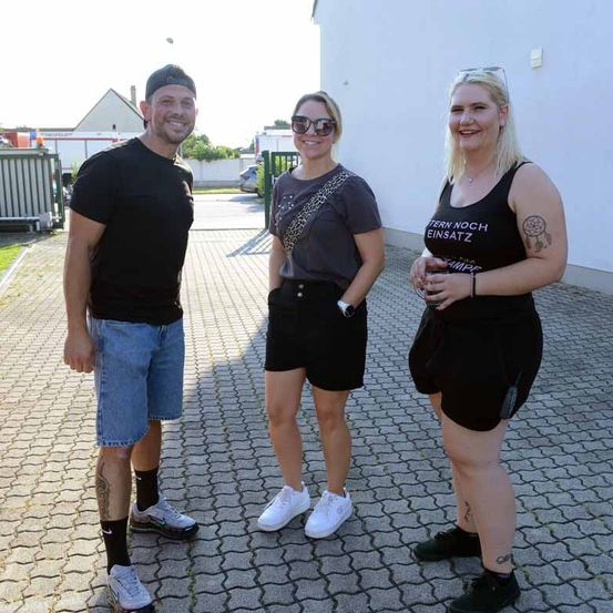 Drei Personen stehen lächelnd vor einem Gebäude. Ein Mann in schwarzen Shorts und einem T-Shirt steht links, eine Frau in schwarzen Shorts und einem grauen T-Shirt steht in der Mitte und eine weitere Frau in schwarzen Shorts und einem schwarzen Tanktop steht rechts.