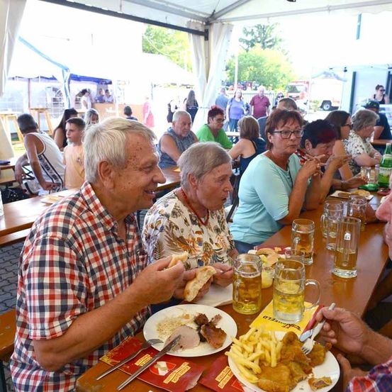 Eine Gruppe von Menschen sitzt an einem Tisch in einem Outdoor-Essbereich. Sie essen und trinken. Der Tisch hat Teller mit Essen, Gläser mit Bier und eine Gabel. Die Menschen lächeln und schauen etwas an. Im unscharfen Hintergrund befinden sich Zelte, Menschen und ein Fahrzeug.