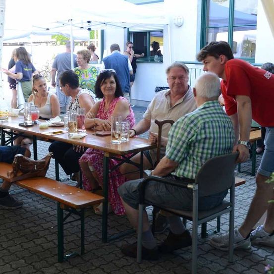 Eine Gruppe von Menschen sitzt an einem Tisch im Freien, einige stehen im Hintergrund. Auf dem Tisch sind Getränke und Essen. Ein Mann hält einen Stock. Ein Mann in Rot lehnt sich über den Tisch.