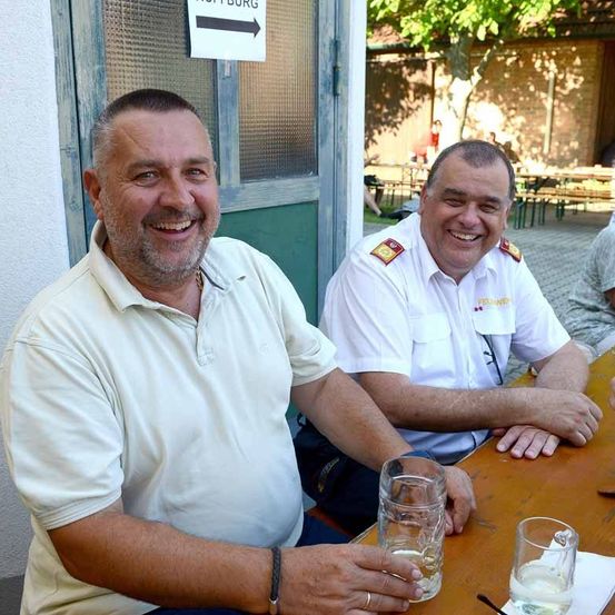 Zwei Männer sitzen an einem Tisch, lächeln und posieren für ein Foto. Ein Mann trägt eine Uhr und hält ein Glas. Der andere Mann ist in Uniform. Sie sitzen in einem Außenbereich. Hinter ihnen ist eine Wand mit einem Schild.