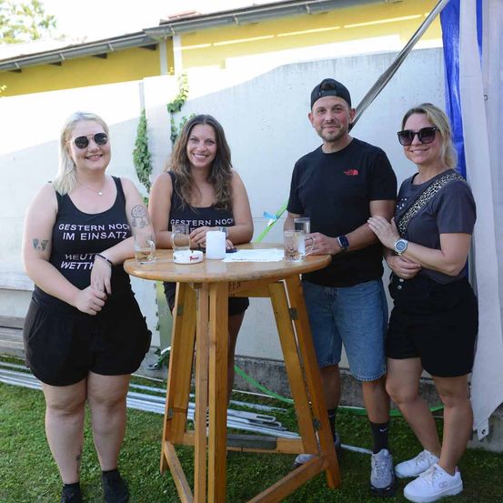 Vier Menschen stehen lächelnd im Freien neben einem kleinen Tisch mit Gläsern und einer Kerze. Zwei Frauen tragen schwarze Tanktops, eine hat eine Tätowierung. Ein Mann in einem schwarzen T-Shirt hält ein Glas. Hinter ihnen befindet sich eine weiße Wand mit Pflanzen.